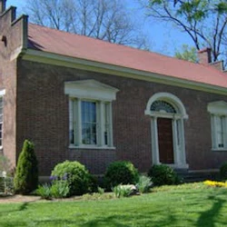 A single-story brick house with a red roof, white-trimmed windows, an arched front door, and a well-kept lawn with shrubs.