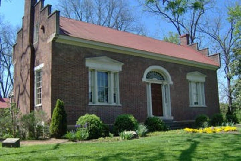 A single-story brick house with a red roof, white-trimmed windows, an arched front door, and a well-kept lawn with shrubs.