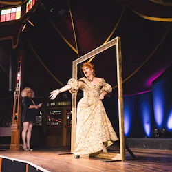 En artist i en utsmyckad klänning poserar dramatiskt inom en stor gyllene ram på scenen; en annan individ står i bakgrunden.