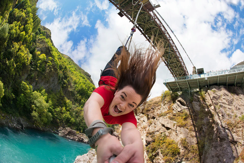 A person in a red shirt bungee jumping from a bridge, with a background of blue sky, greenery, and water below.