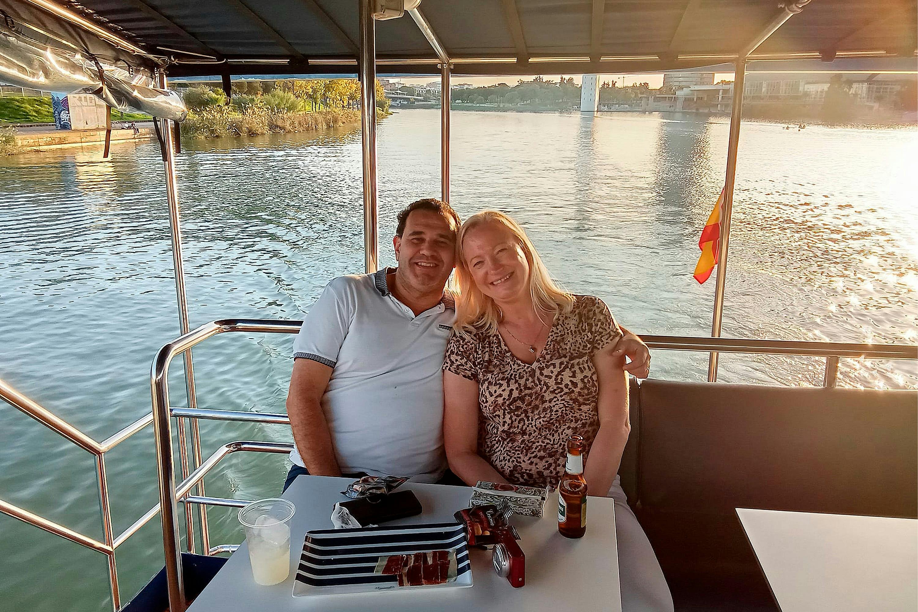 Een lachend stel zittend aan een tafel op een boot, met eten en drinken, met uitzicht op een kalme rivier en een stadsgezicht op de achtergrond.