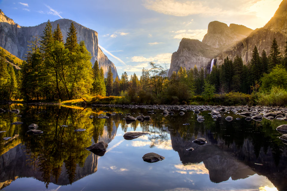 Mountainous landscape with a waterfall, trees, and rocks, all reflected in a tranquil river under a partially cloudy sky at sunset.