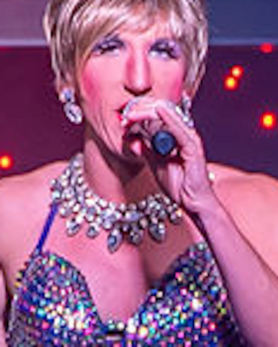Performers in sparkly outfits singing on stage, with colorful lights and an audience visible in the foreground.
