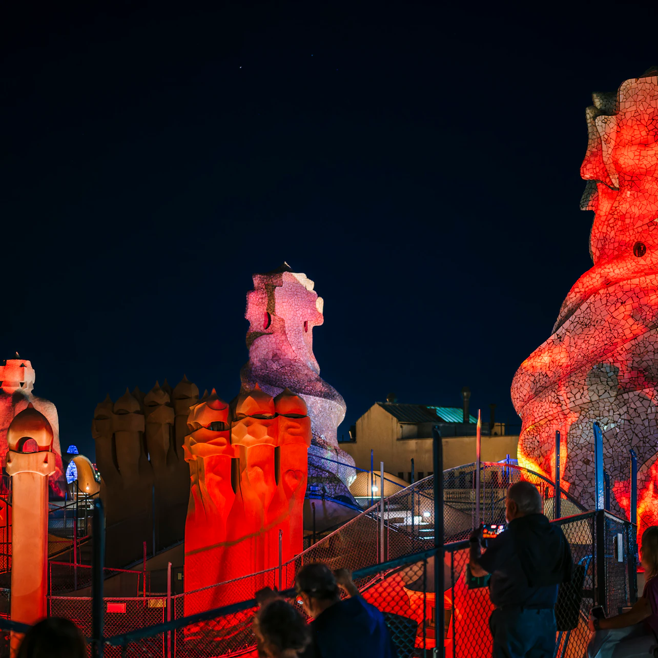 Tourists on a rooftop at night admire colorful, illuminated, abstract stone sculptures at a famous architectural site.