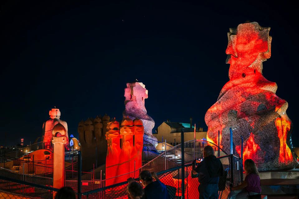 Tourists on a rooftop at night admire colorful, illuminated, abstract stone sculptures at a famous architectural site.