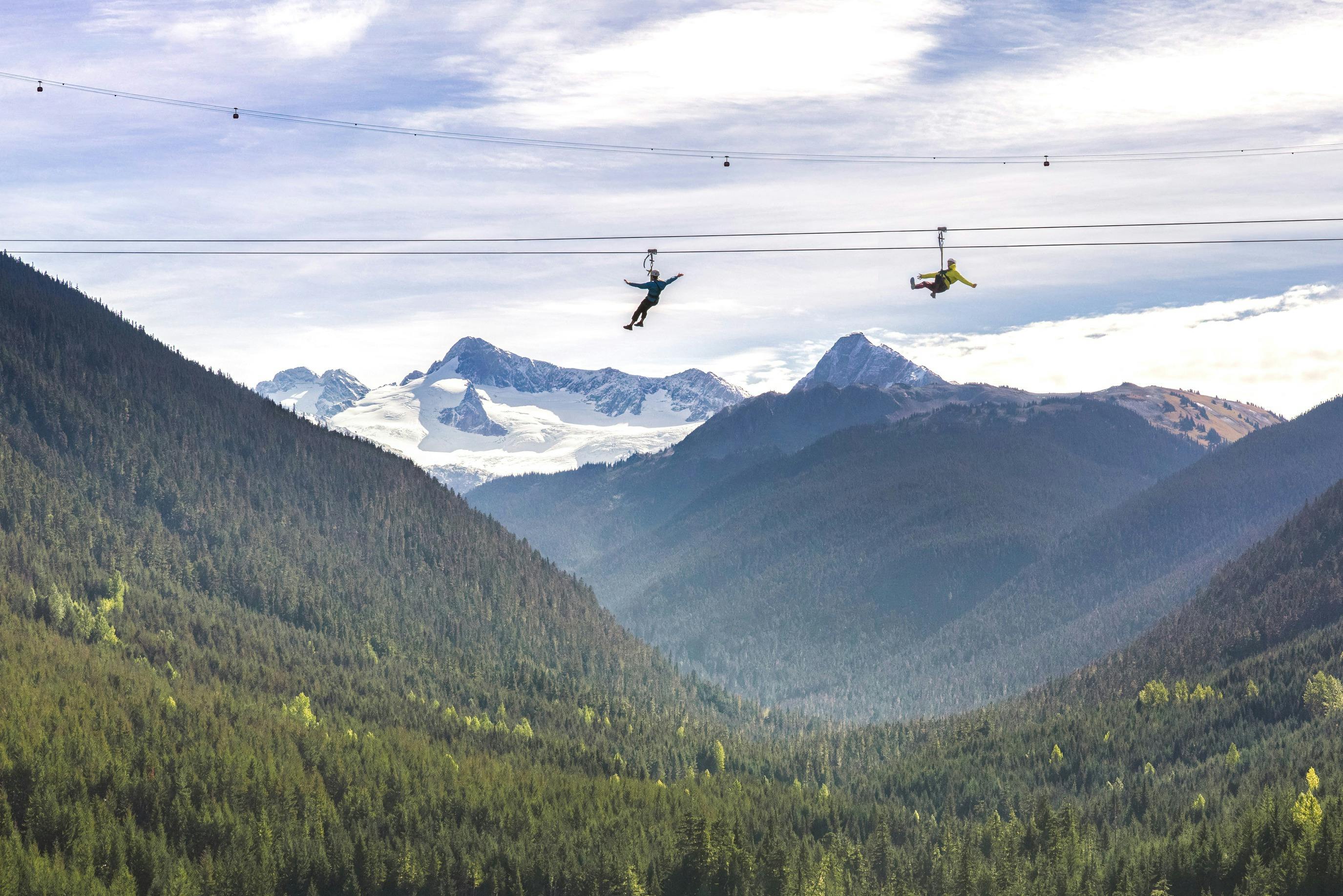 부분적으로 흐린 하늘 아래 눈 덮인 산을 배경으로 울창한 숲을 가로지르는 두 사람.