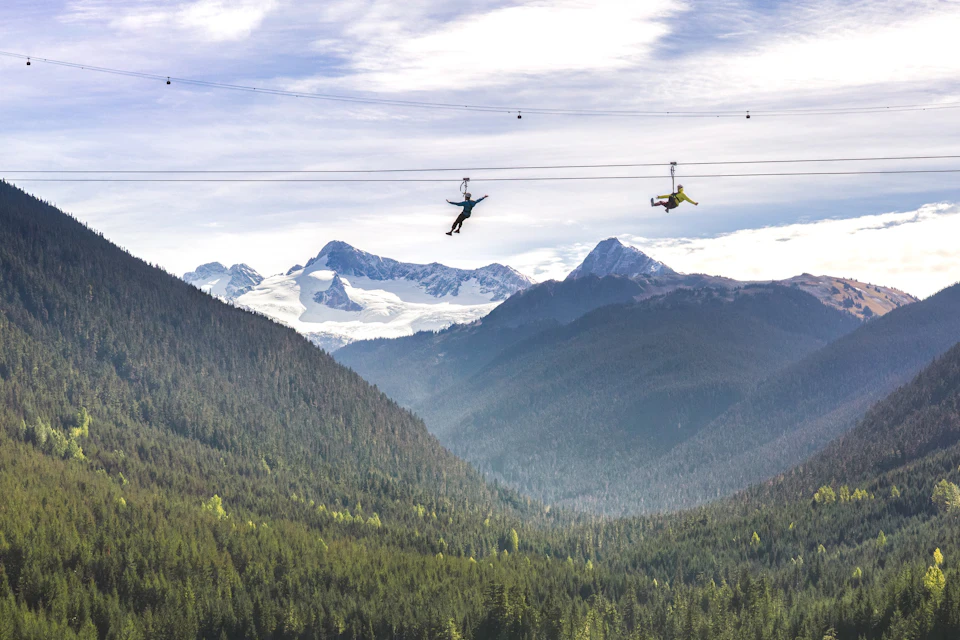 Two people ziplining over a lush forest with snow-capped mountains in the background under a partly cloudy sky.