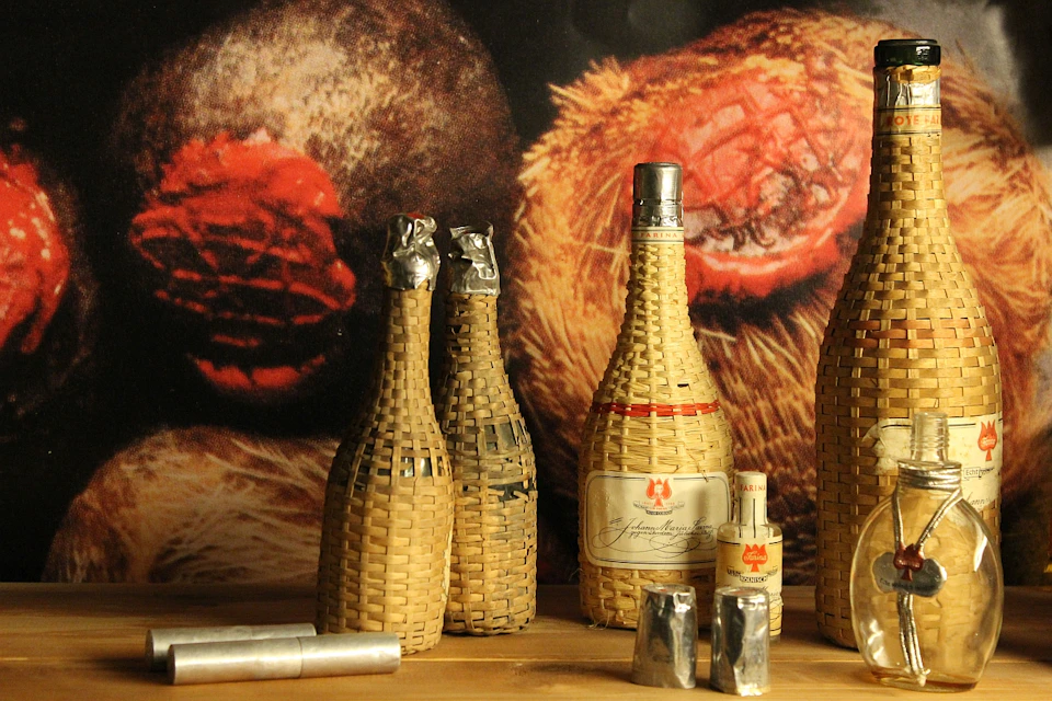 Four wicker-covered bottles and two metal cylinders on a wooden surface, with a textured fabric background.