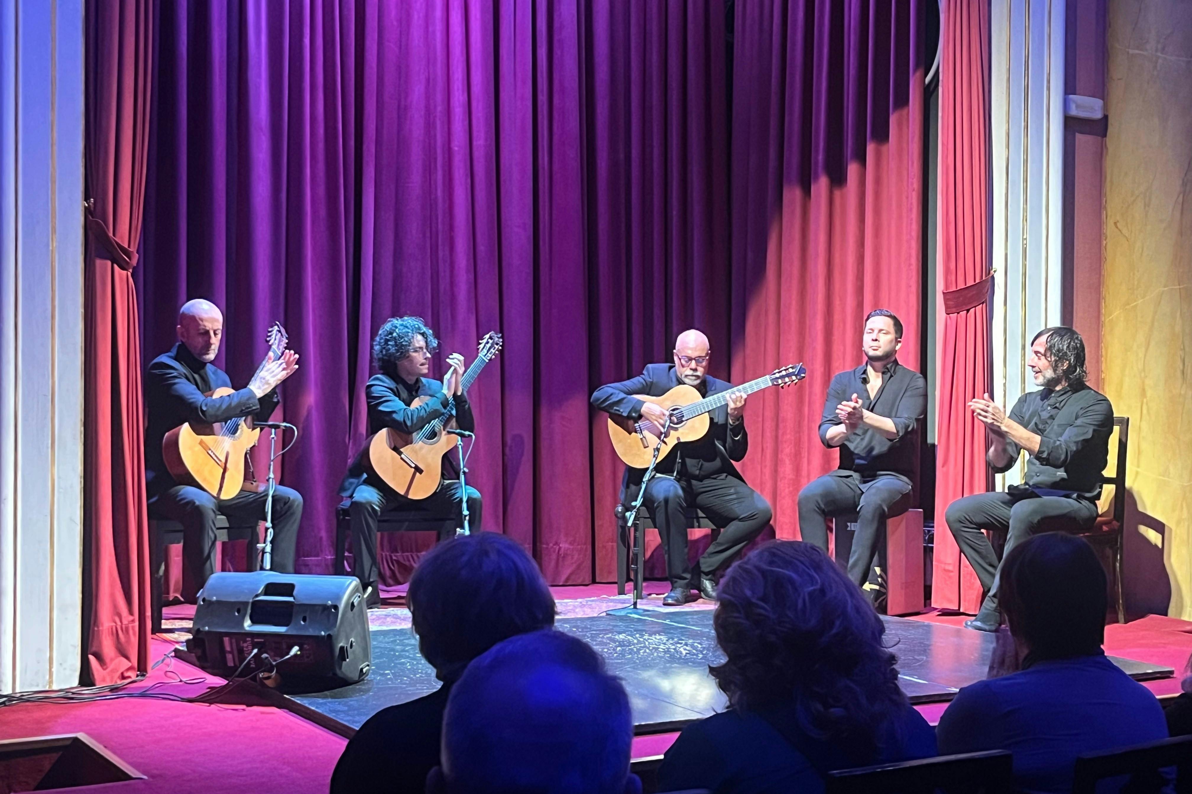 Quatre musiciens se produisent sur scène avec des guitares et des applaudissements, assis devant des rideaux rouges, sous le regard d'un public.
