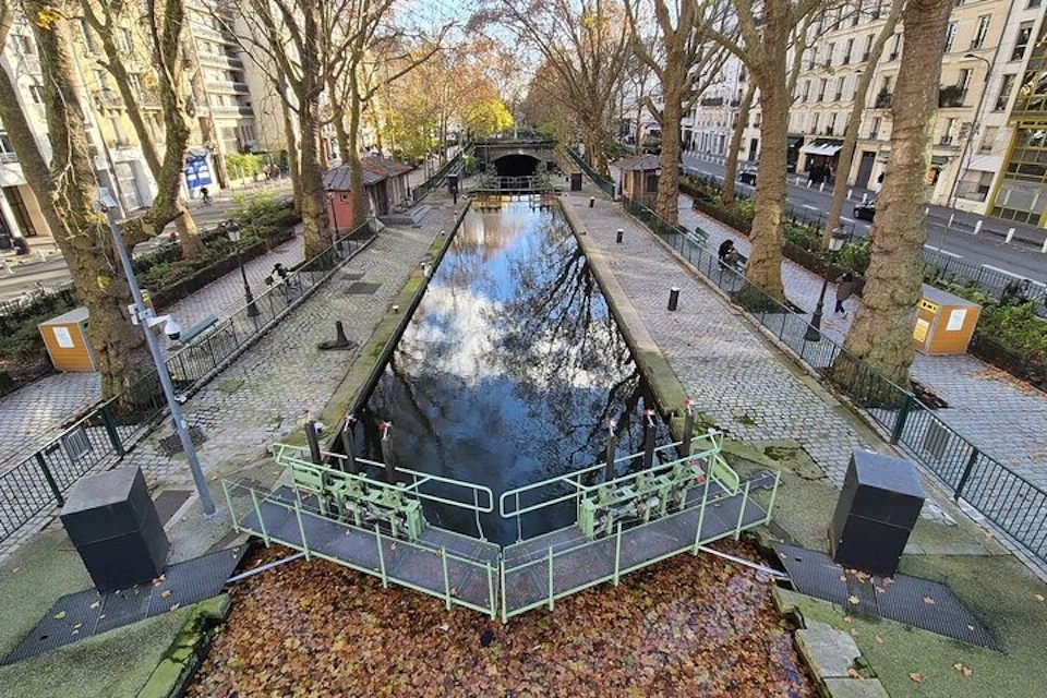 A canal with reflective water, flanked by cobblestone paths and trees, in a cityscape under a partly cloudy sky.
