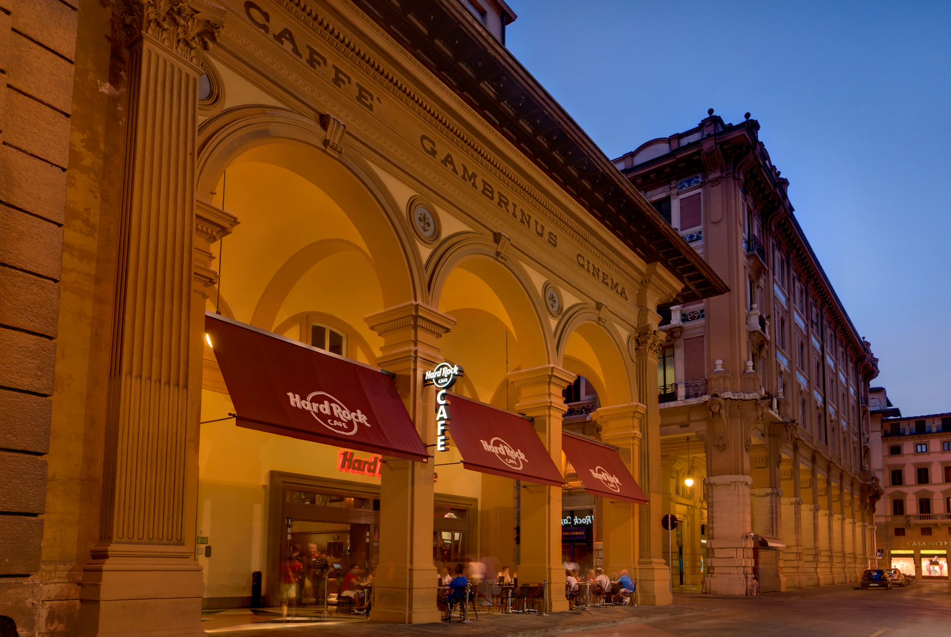 An illuminated café with arches and "Caffè Gambrinus Cinema" signage. People seated under red "Hard Rock Café" awnings.