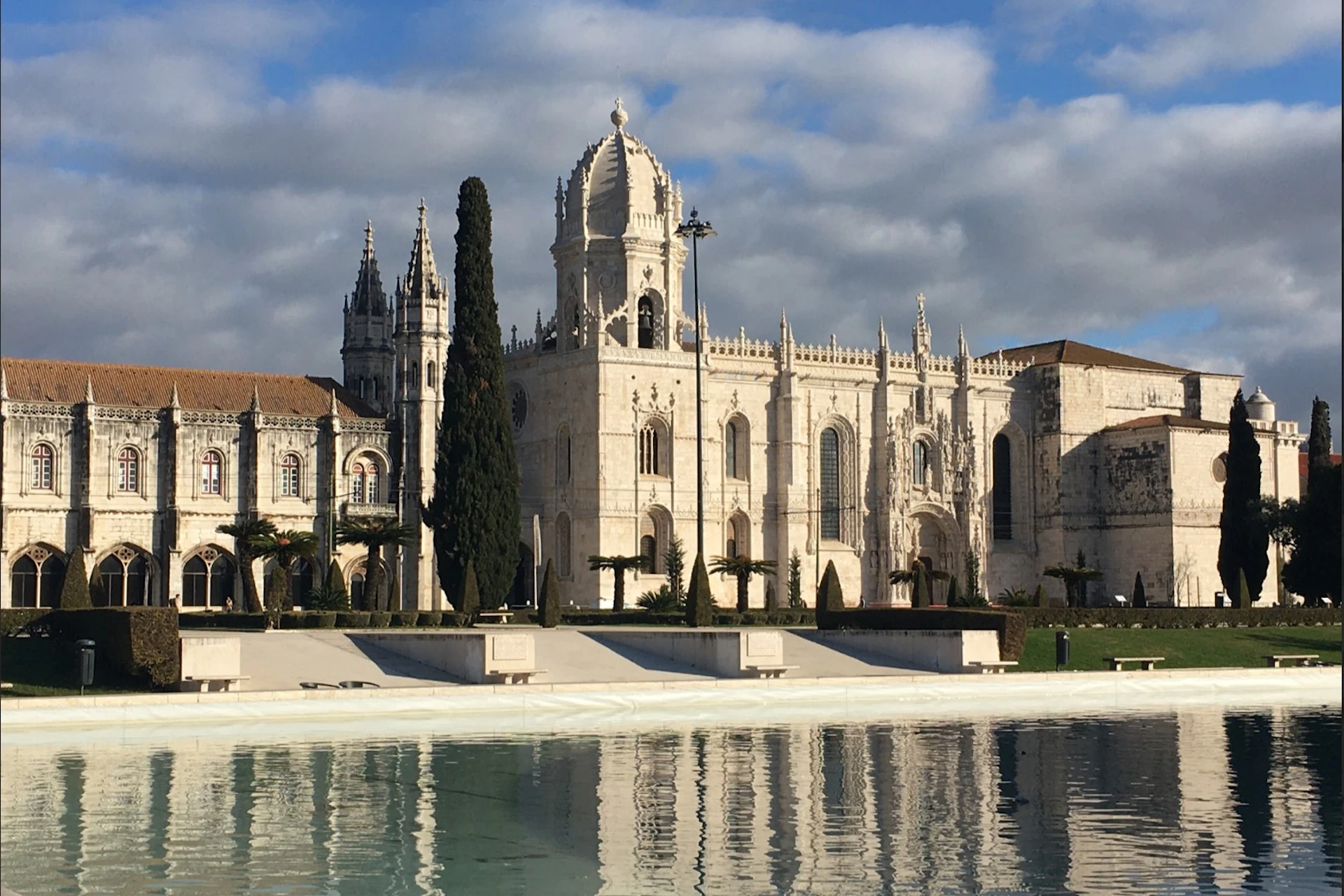 A grand, white historic building with ornate architecture stands behind a reflecting pool and tall trees under a cloudy sky.