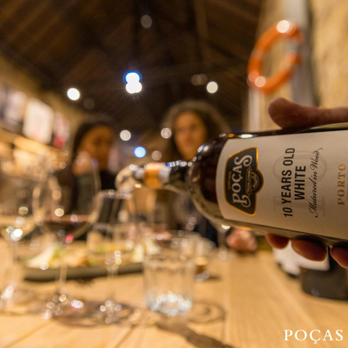 Una botella con la etiqueta "Poças 10 Años Oporto Blanco" siendo vertida en un vaso sobre una mesa de madera con gente al fondo.