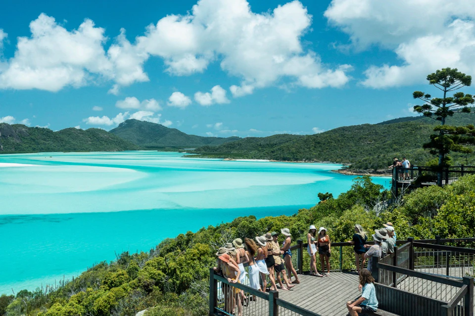 Group of people on a viewing platform overlooking a scenic turquoise bay surrounded by lush greenery and hills under a partly cloudy sky.