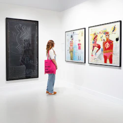 Person med en rosa väska som läser "In art we trust" som observerar inramade konstverk på en gallerivägg.