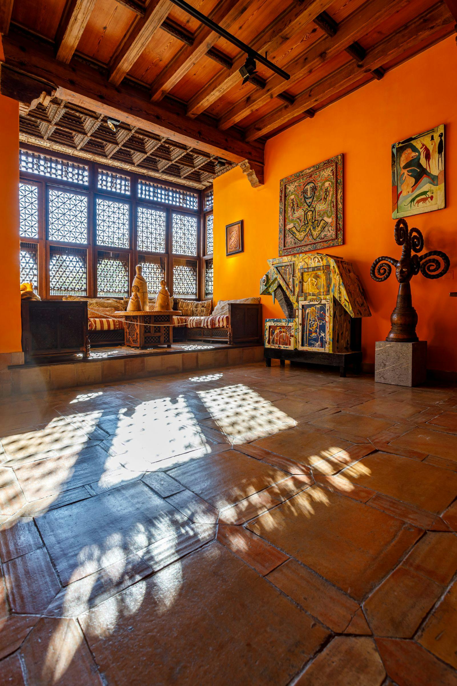 A vividly lit room with orange walls, wooden furniture, patterned windows, eclectic decor, and sunlight casting intricate shadows.
