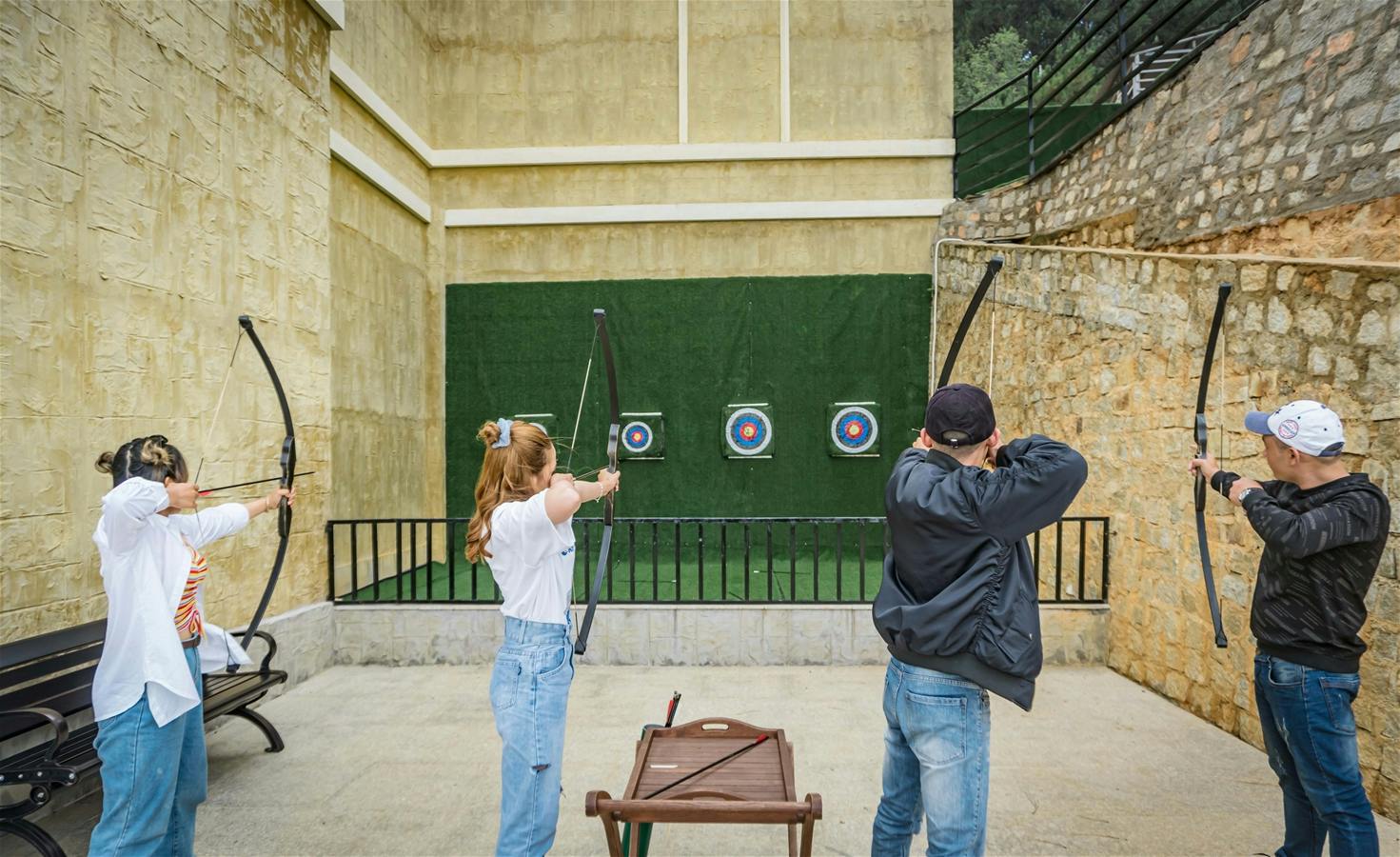 Three people practice archery, aiming at targets on a green wall in an outdoor setting with beige walls and fencing.