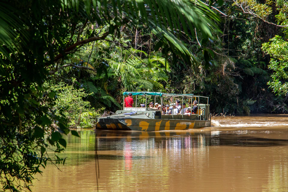 A safari-style boat with people aboard cruising on a river surrounded by lush tropical vegetation.