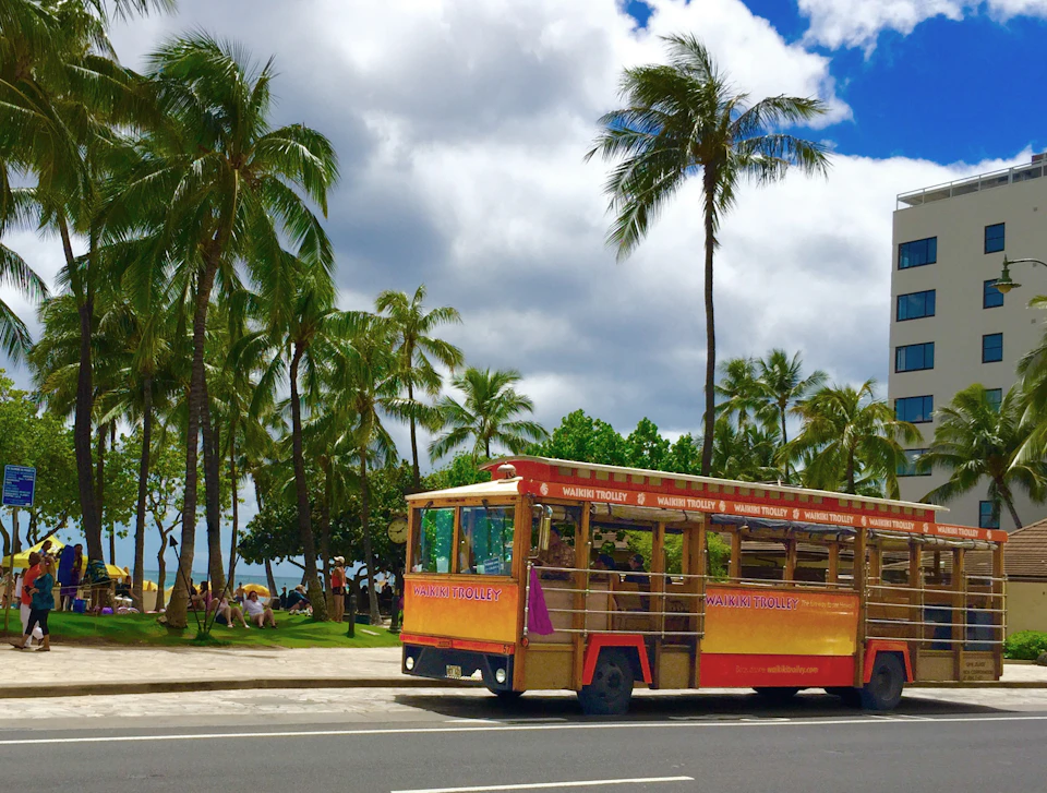 A red and yellow open-air trolley labeled "Waikiki Trolley" on a road with palm trees and people sitting in the background.