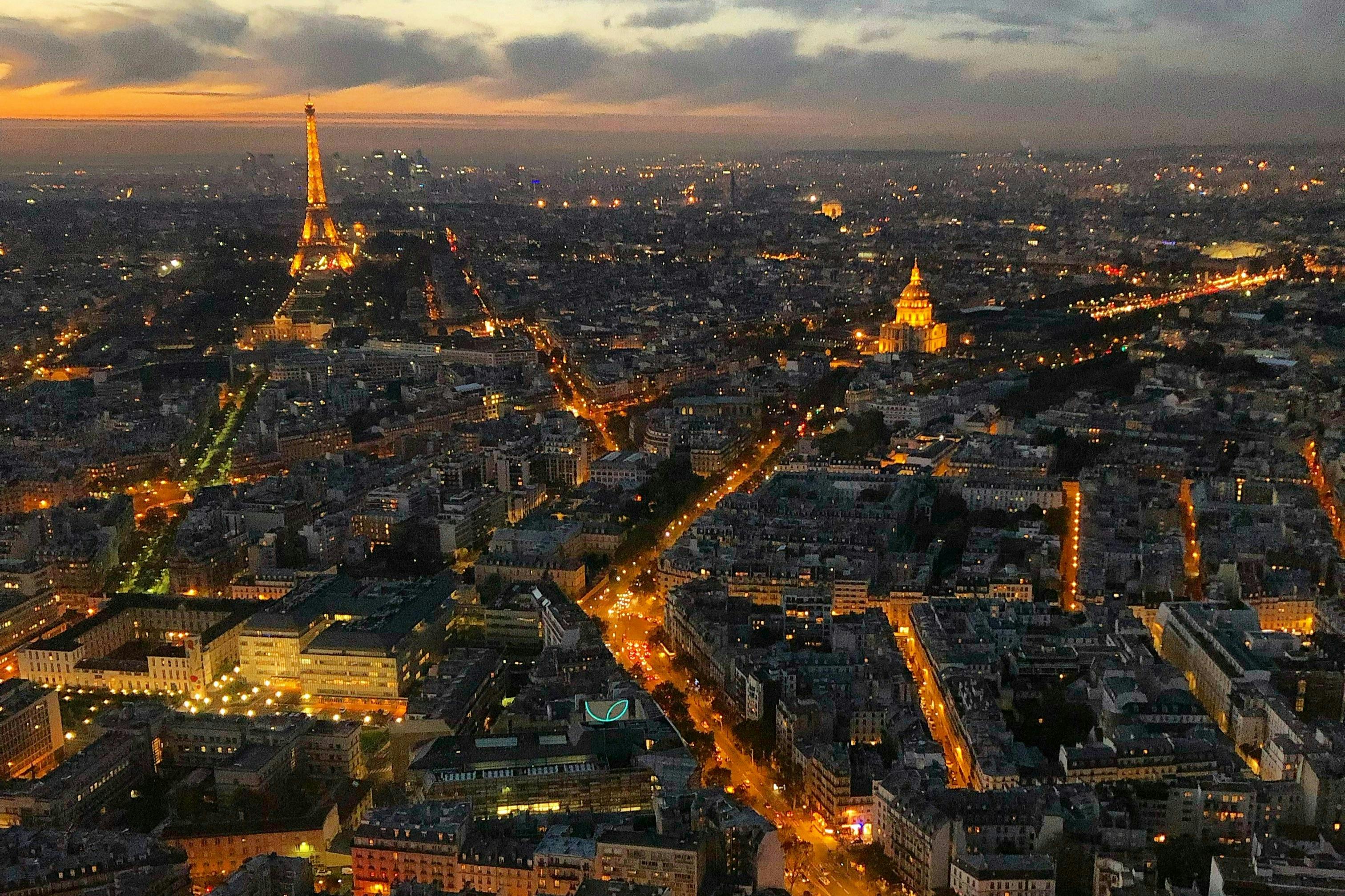 Aerial view of Paris at dusk, featuring the Eiffel Tower and the dome of Les Invalides, with illuminated streets and buildings.