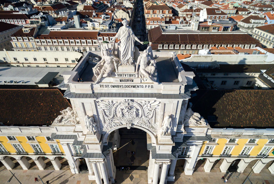 Aerial view of an ornate archway in a city with detailed sculptures on top and buildings lining a street behind it.