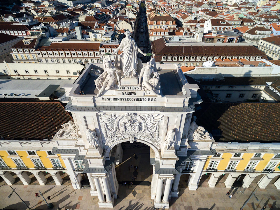 Aerial view of an ornate archway in a city with detailed sculptures on top and buildings lining a street behind it.
