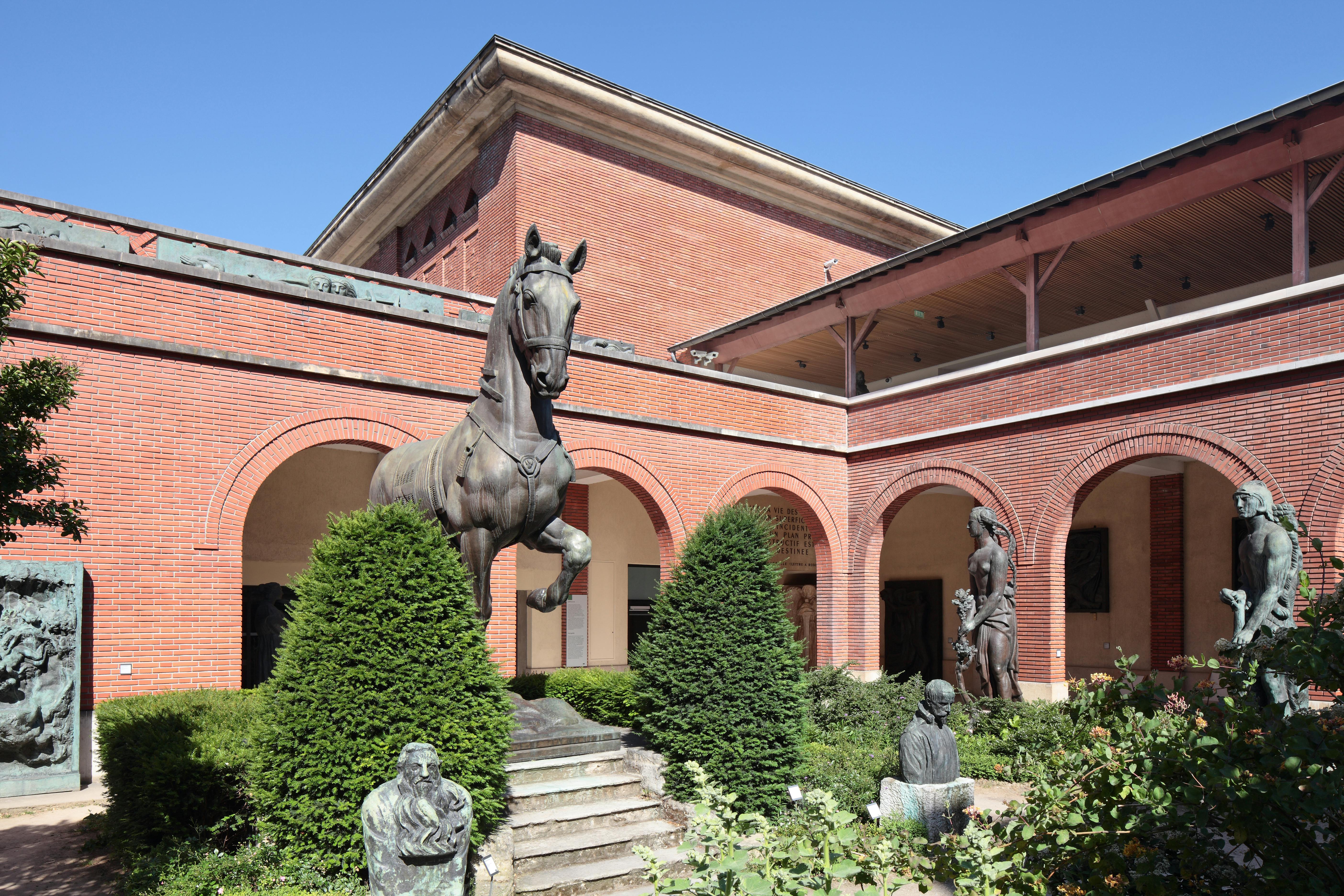 A courtyard with red brick walls, featuring bronze sculptures of a horse, human figures, and lush greenery.