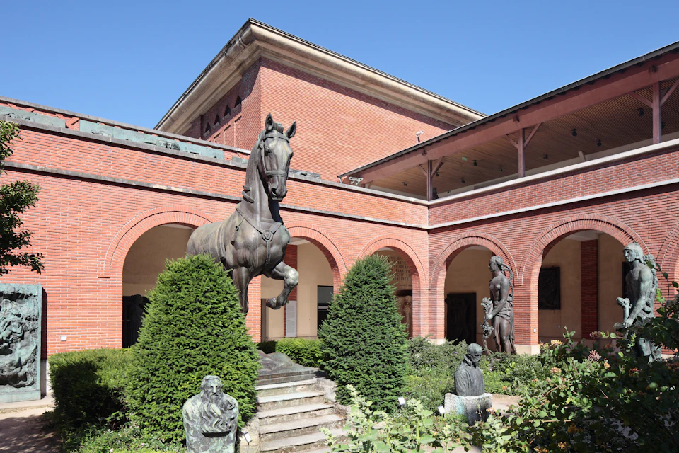 A courtyard with red brick walls, featuring bronze sculptures of a horse, human figures, and lush greenery.