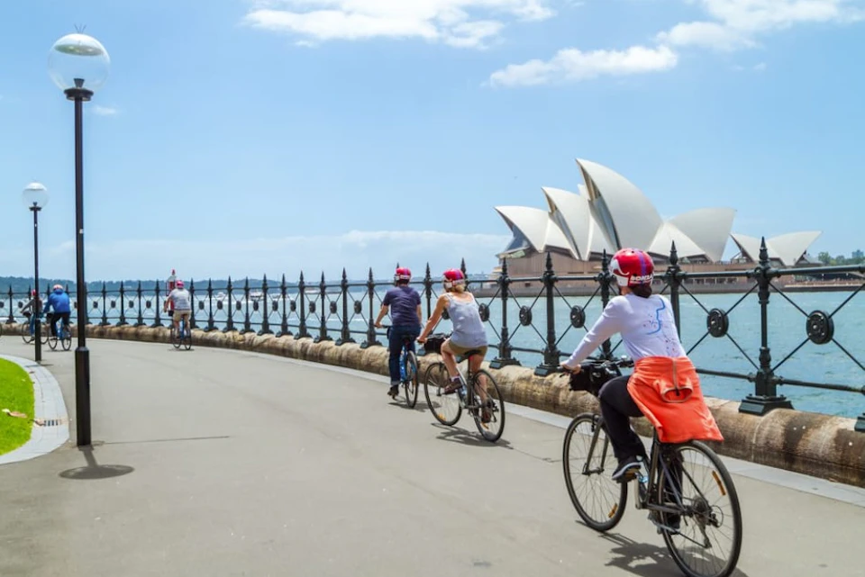 Cyclists riding along a waterfront path with the Sydney Opera House visible in the background under a clear sky.