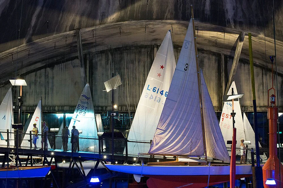 People viewing sailboats displayed in an indoor exhibit with dramatic lighting.