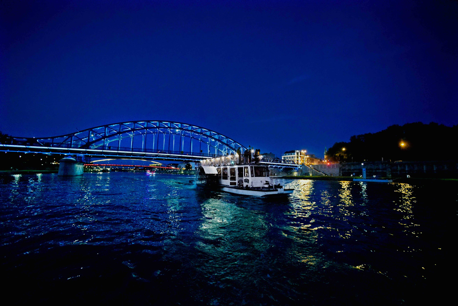 A boat on a river at night under illuminated blue bridges with city lights reflecting on the water.