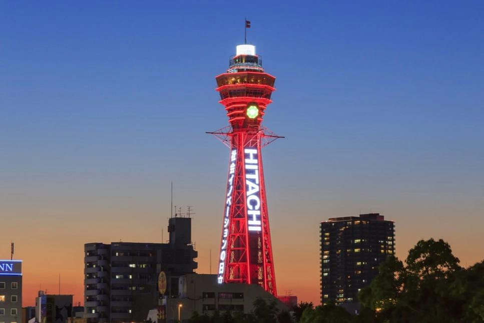 A red-lit observation tower with the word 'Hitachi' displayed, set against a twilight sky and surrounded by buildings.