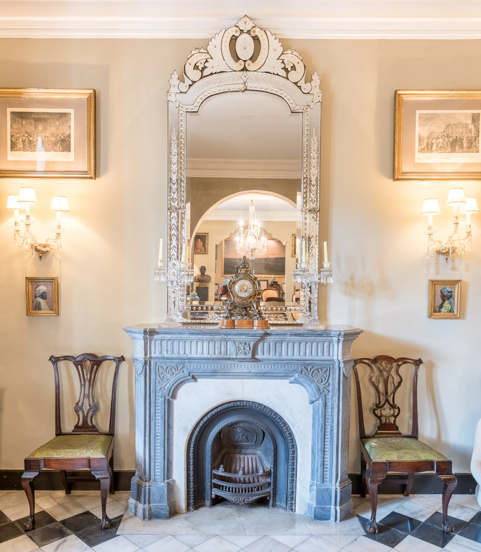 Elegant room with ornate fireplace, large mirror, two chairs, wall sconces, and framed artwork, reflecting a chandelier.