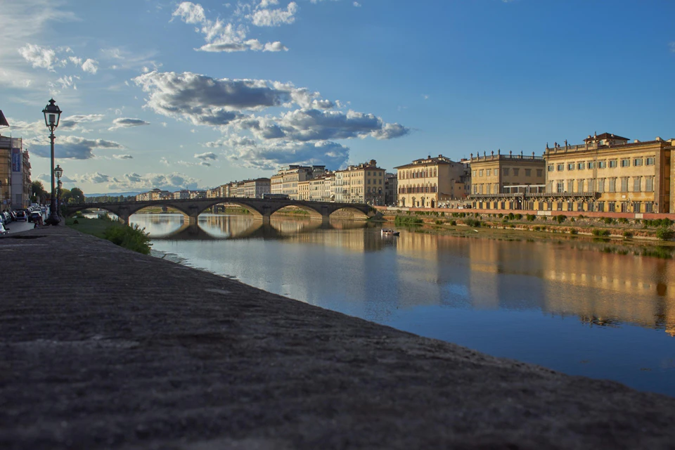A calm river with bridges and historic buildings along the banks under a blue sky with scattered clouds.