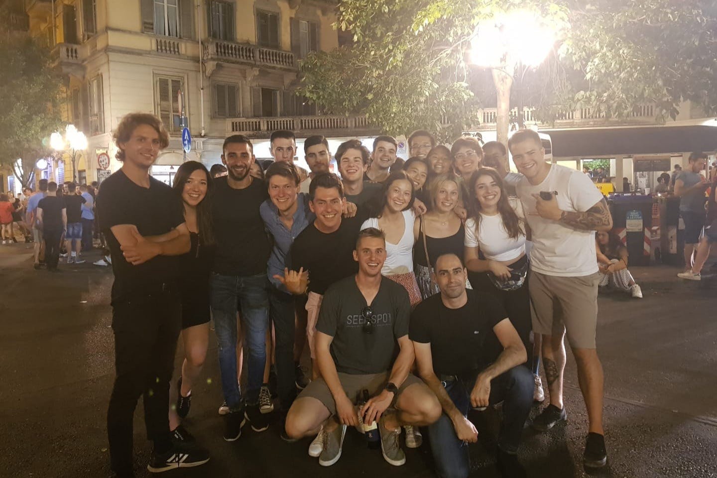 A group of people smiling and posing for a photo on a street at night, with a building and trees in the background.