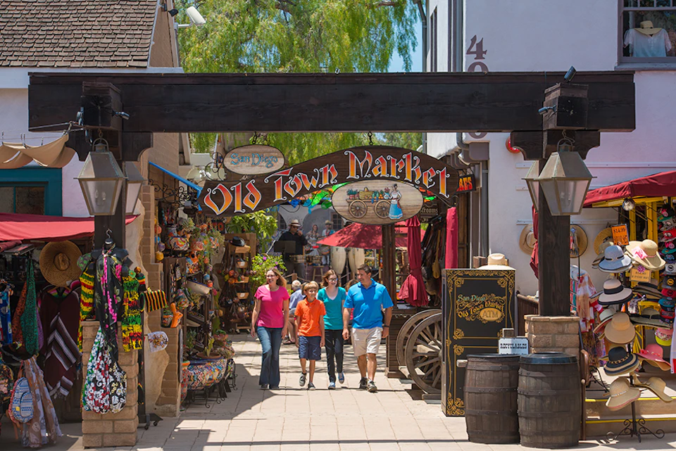 Group of people walking under "Old Town Market" sign in a market area with shops, colorful items, a wagon, and barrels.