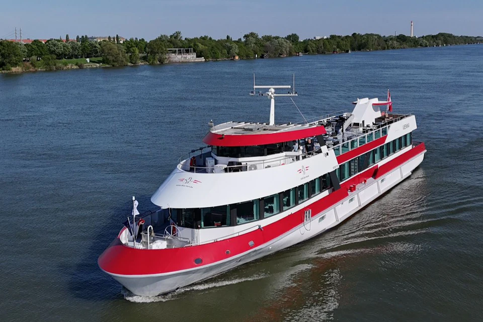A red and white passenger boat cruises on a river, with greenery and buildings visible along the distant shoreline.