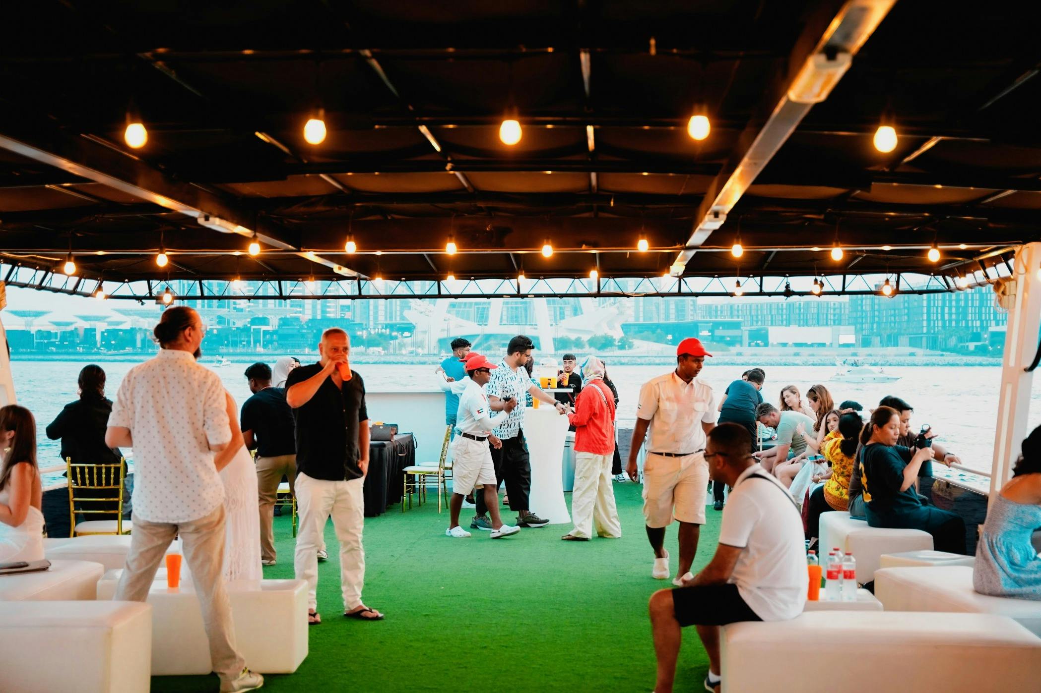 Människor som umgås på ett båtdäck med konstgräsgolv, strängljus över huvudet och en stadsbild vid vattnet i bakgrunden.