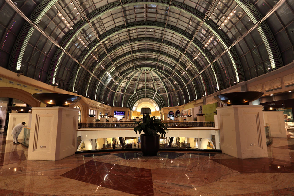 Spacious indoor mall with a grand arched glass ceiling, marble floors, and a large potted plant in the center.
