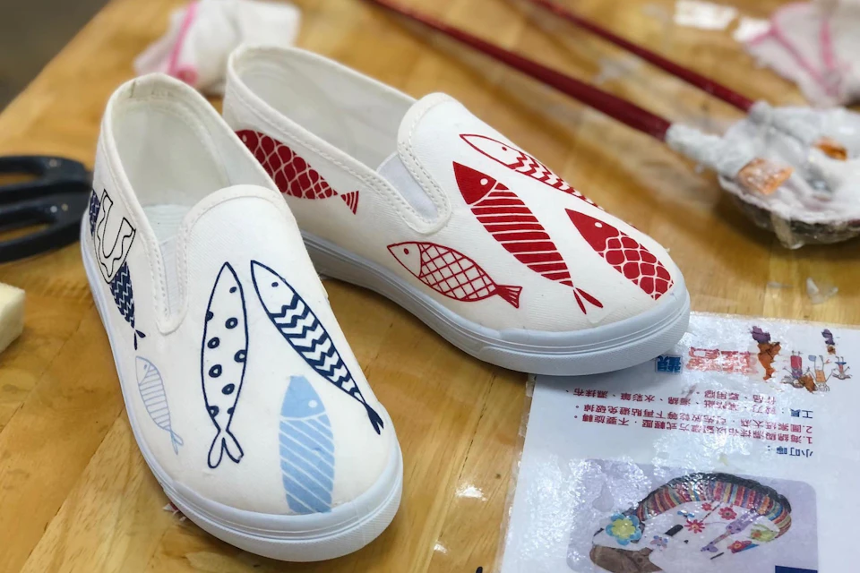 White slip-on shoes with red and blue fish designs on a wooden table, accompanied by art supplies.