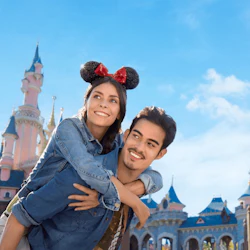 En leende kvinna som bär Mimmi Pigg-öron rider på en mans rygg framför ett sagoslott under en blå himmel.