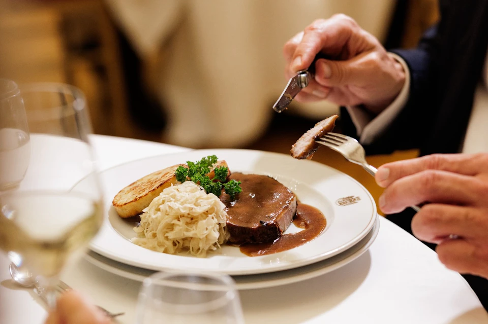 A person cutting into a steak on a white plate with mashed potatoes, sauerkraut, and a garnish, seated at a dining table.