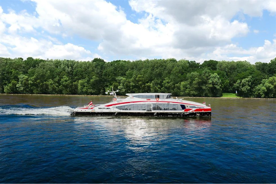 Modern passenger ferry with red and white design cruising past a tree-lined shore under a partly cloudy sky.