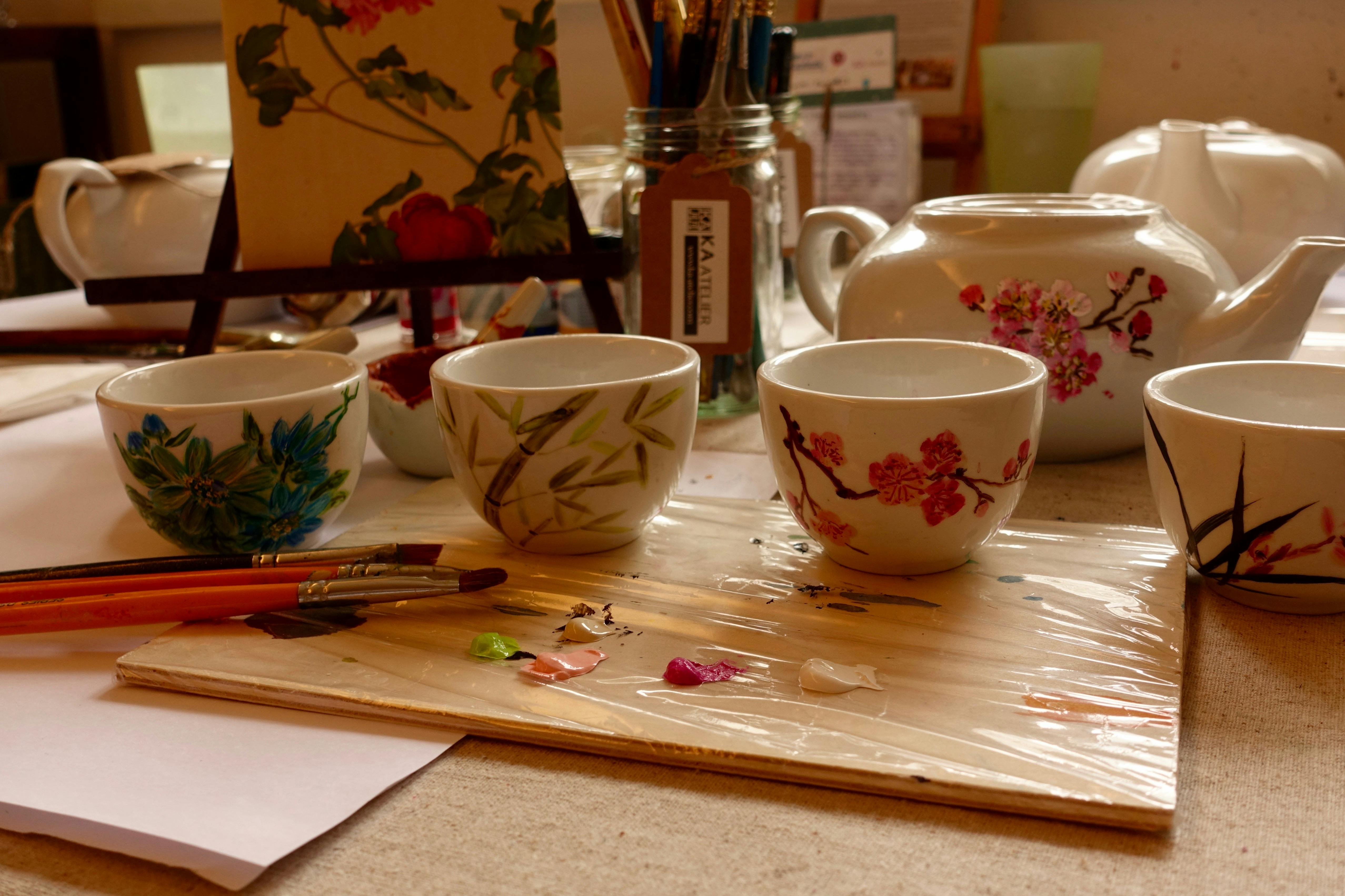 Three painted teacups and paintbrushes on a table with a teapot and painting supplies in the background.