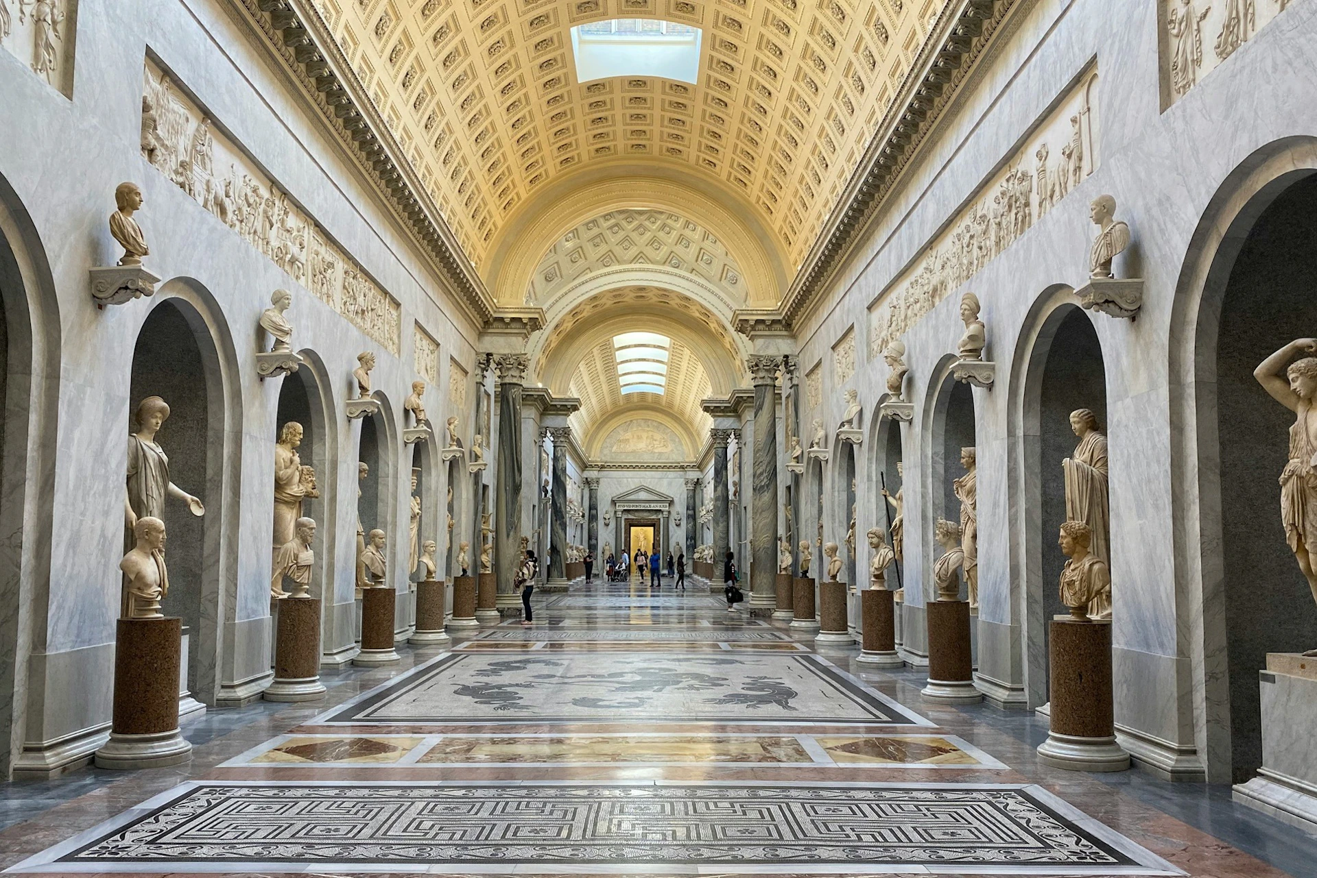 A grand hall with ornate arched ceilings, statues in niches along the walls, and a patterned floor, in a museum setting.