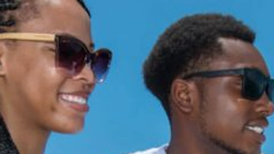 Two people wearing sunglasses on a boat, laughing and looking out to sea under a clear, blue sky.