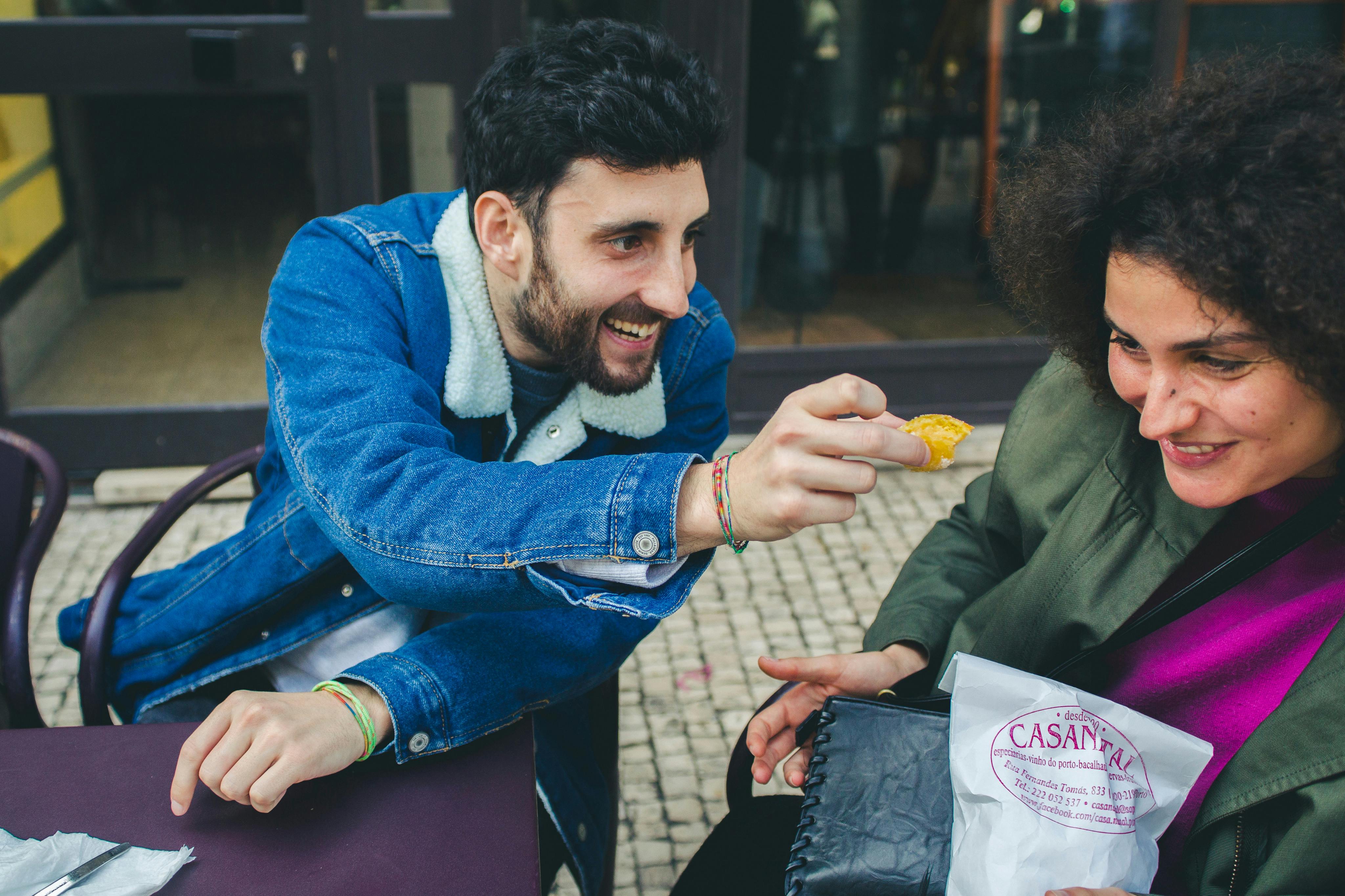 Ένας άντρας χαμογελά ενώ προσφέρει φαγητό σε μια καθισμένη γυναίκα που κρατά μια λευκή χάρτινη τσάντα με κόκκινο κείμενο σε εξωτερικούς χώρους σε ένα πλακόστρωτο πεζοδρόμιο.