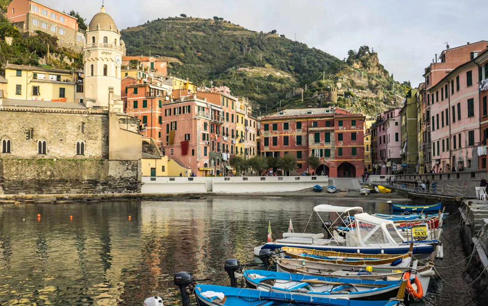Colourful buildings line a calm waterfront with boats docked in the foreground, and a green hillside in the background.