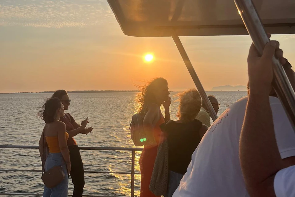 People stand on a boat's deck at sunset, with the sun low in the sky and a calm sea in the background.