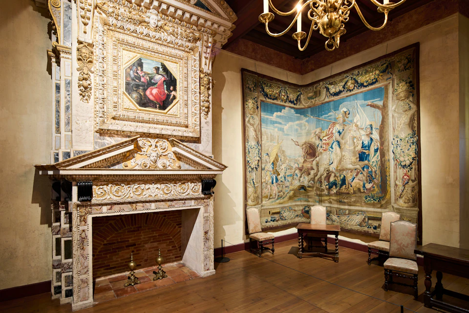 Elegant room with ornate fireplace, framed painting, large tapestry depicting a historical scene, wooden chairs, and a chandelier.